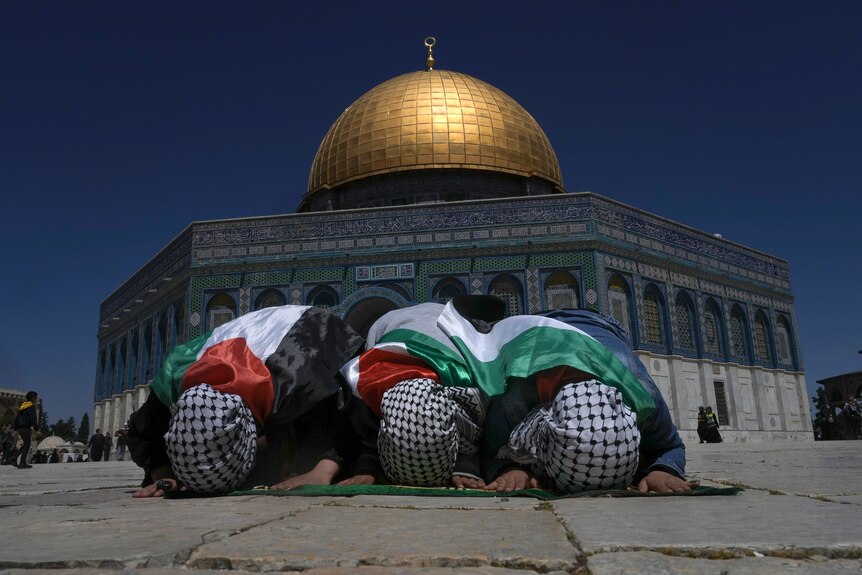 Muslim worshippers wrapped in Palestinian flags pray with heads down with gold dome of mosque in background.
