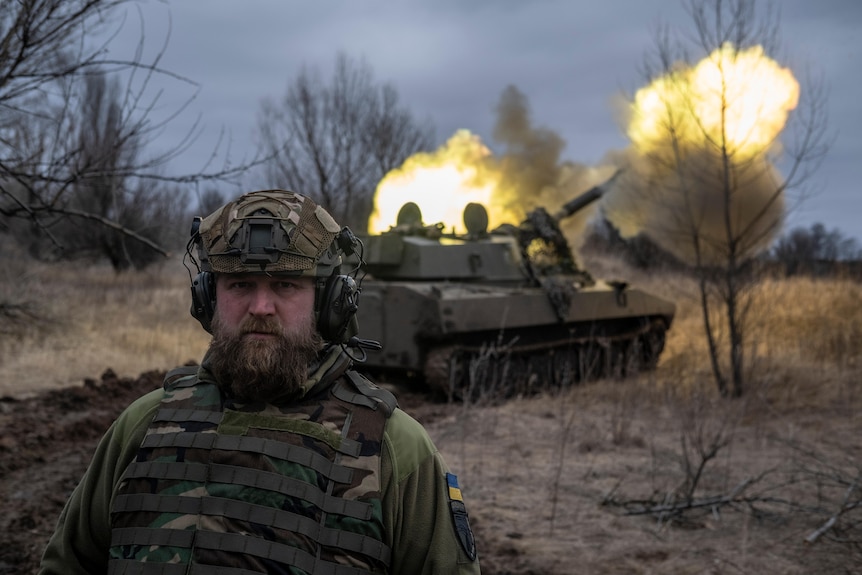 A bearded man in combat gear looks at the camere as a tank fires behind him.