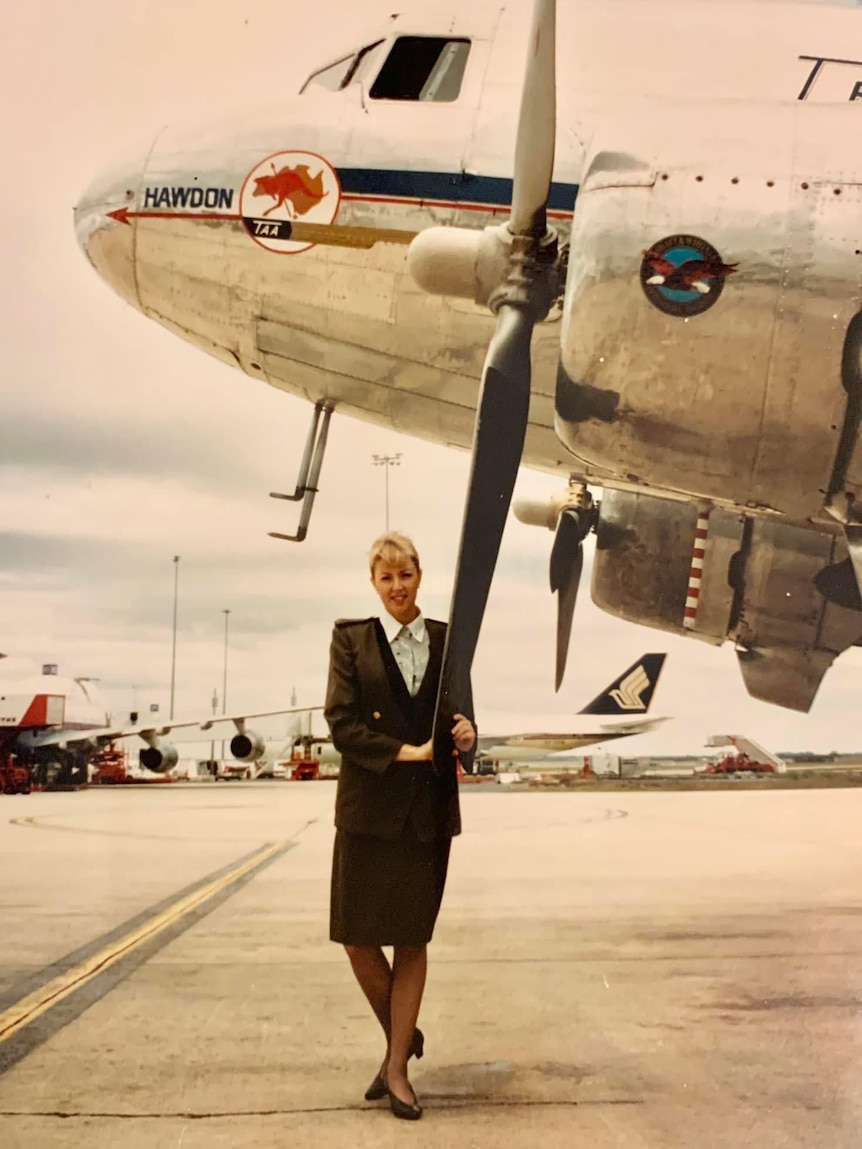 An old faded photo of Louise in her flight attendant uniform standing under a plane and holding the propeller.