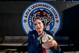 An old man holds a soccer ball in front of an APIA Leichardt logo.
