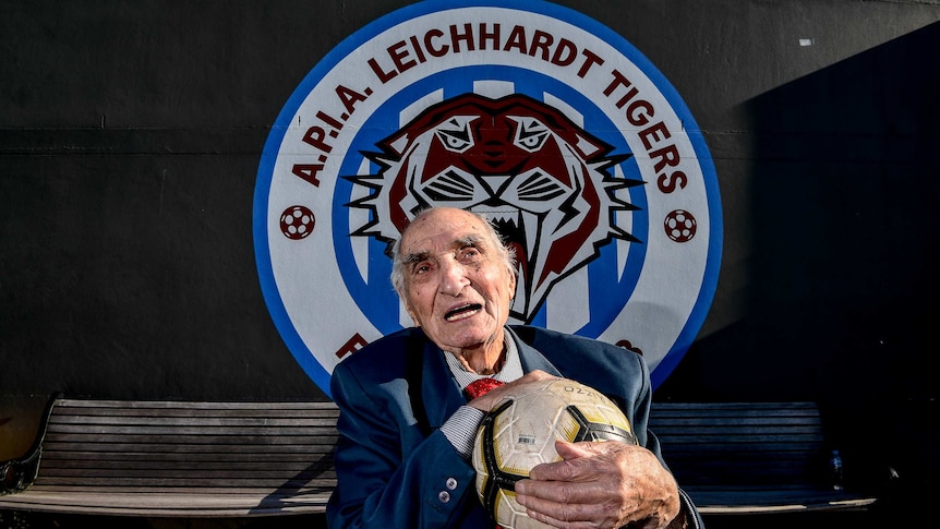 An old man holds a soccer ball in front of an APIA Leichardt logo.