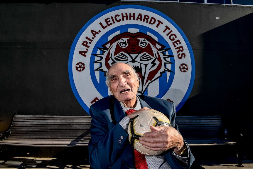 An old man holds a soccer ball in front of an APIA Leichardt logo.