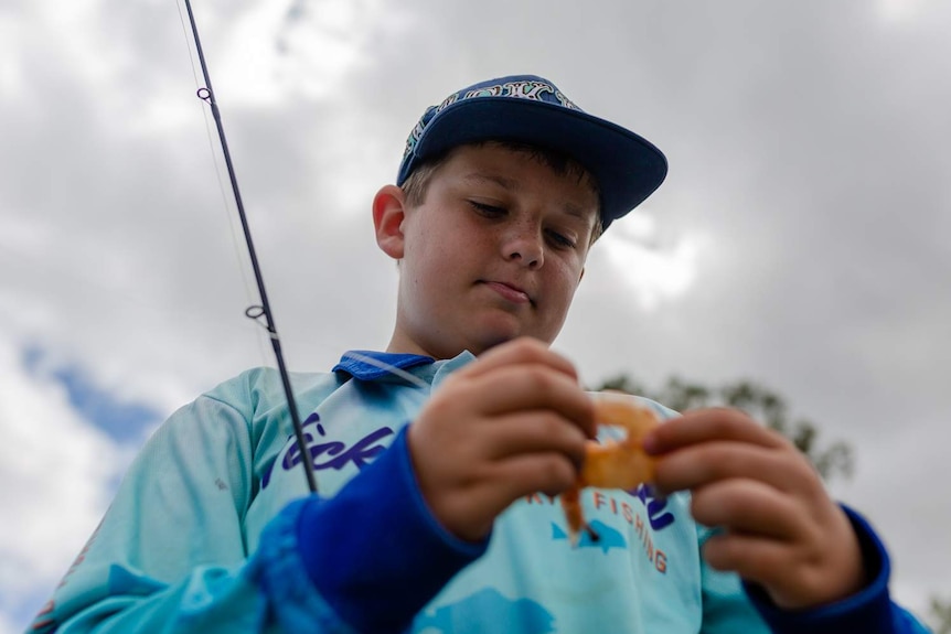 Looking up towards a young boy wearing a cap and holding a fishing rod. Holding prawn in foreground.