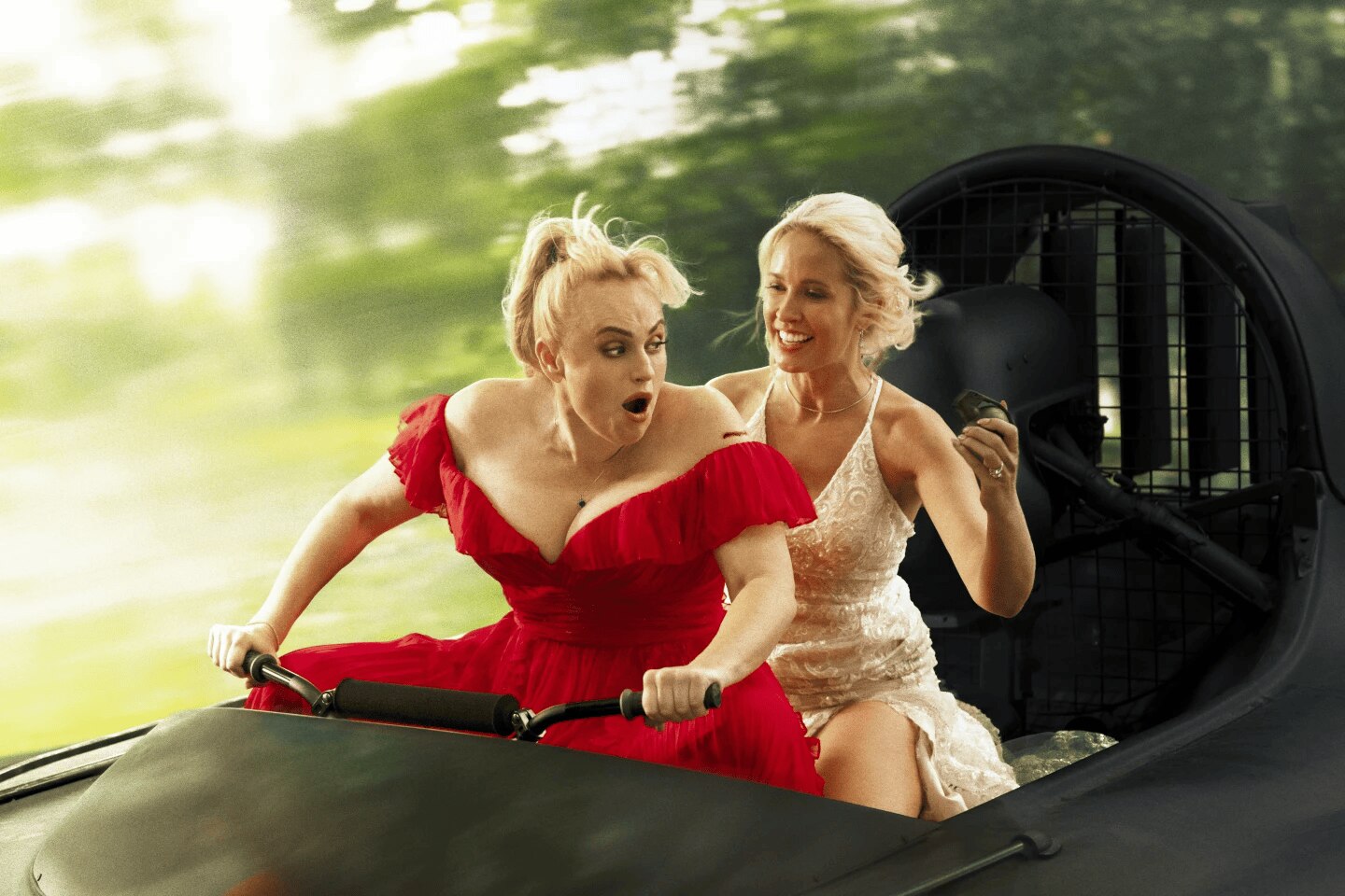 A film still showing a woman wearing a red dress riding a vehicle, a woman in a white dress sitting behind her