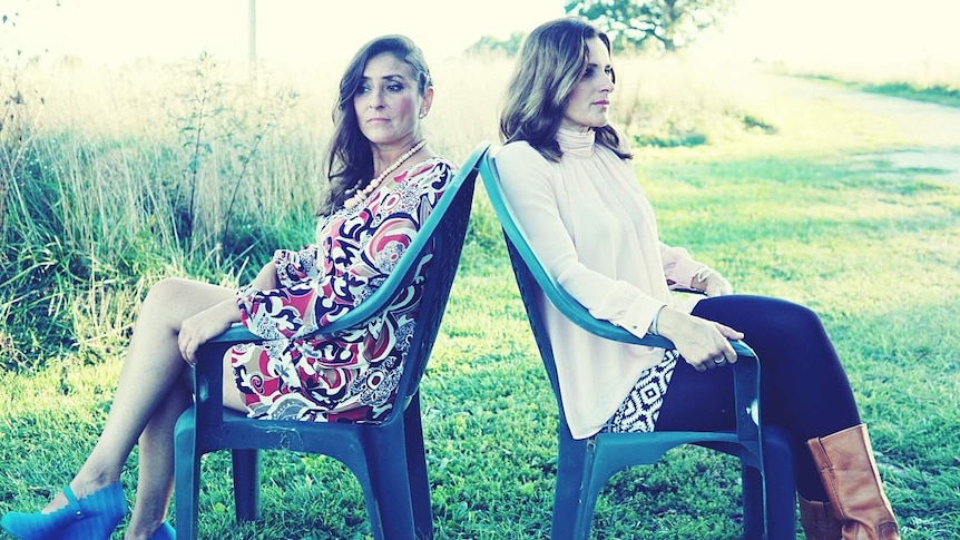 two women sitting outdoors on chairs back to back. One wearing a colourful dress and the other in white blouse and pants