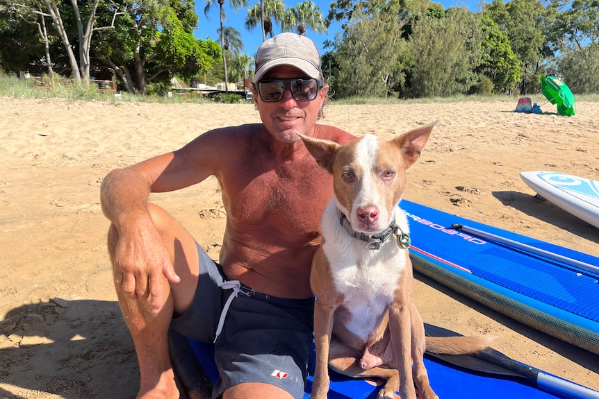 A smiling man, wearing cap, glasses, sits shirtless on a paddleboard with his dog next to him.