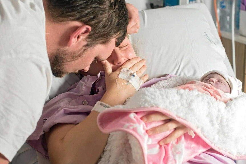 A man and woman cry as the woman holds a stillborn baby wrapped in pink blankets.
