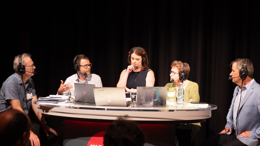 A group of five people sit around a semi-circular table with headphones on