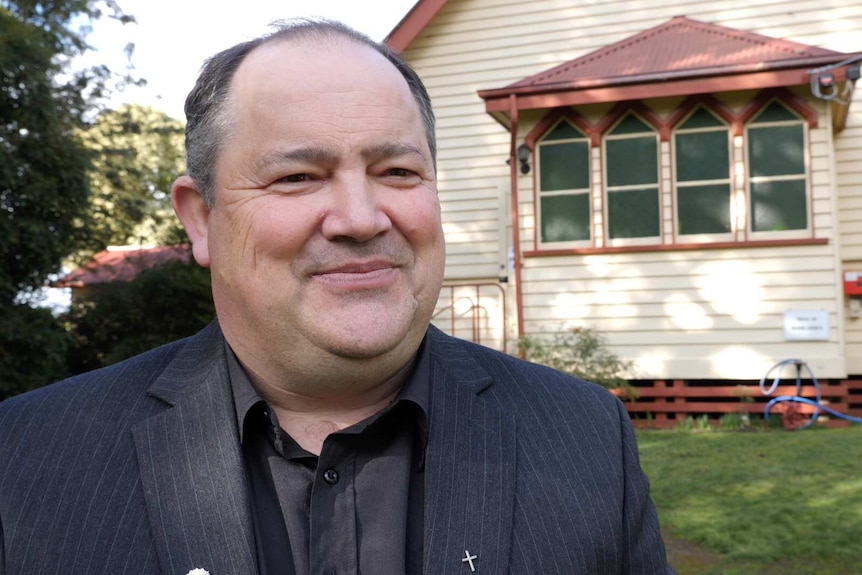 A smiling man in a black shirt and pinstriped jacket stands out the front of an old timber country church.