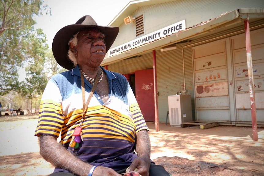 An older Indigenous man wearing an akubra sitting out the front of a community hall in a remote community.