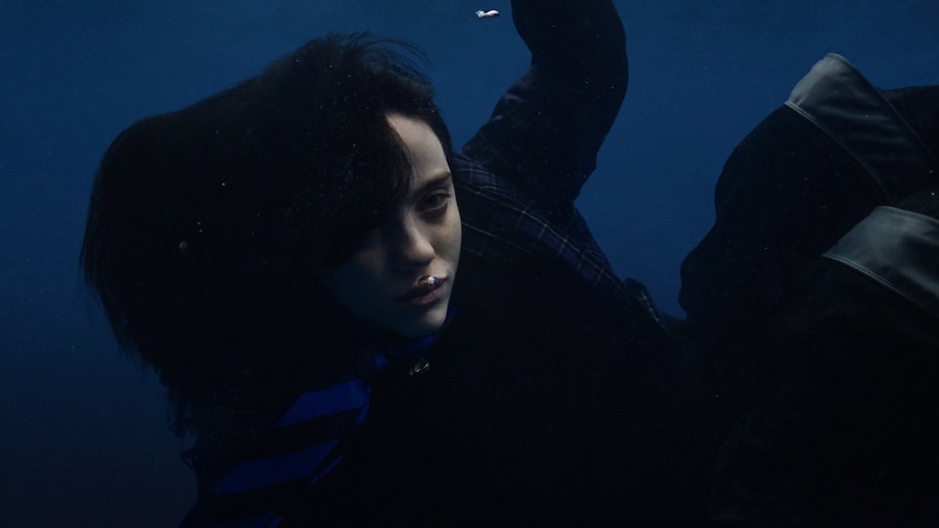 Billie Eilish floats underwater blowing bubbles in a full black, tartan wardrobe
