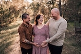 Two men stand on either side of a pregnant woman holding her belly. 