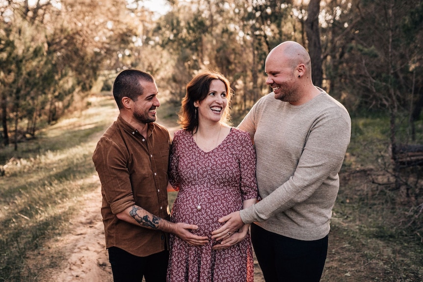 Two men stand on either side of a pregnant woman holding her belly. 