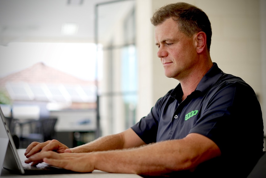 A man wearing a black polo shirt with a green logo typing on a laptop at a desk.