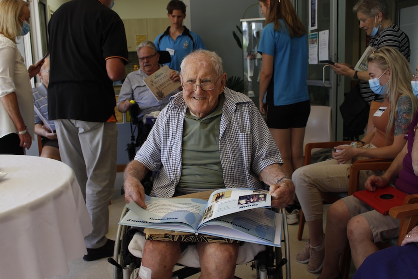 An elderly man in a wheelchair sits smiling, looking at a book.