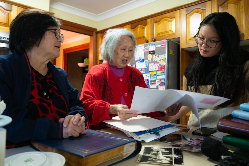 Three women of three generations stand inside a home and look at piles of paper on a table.