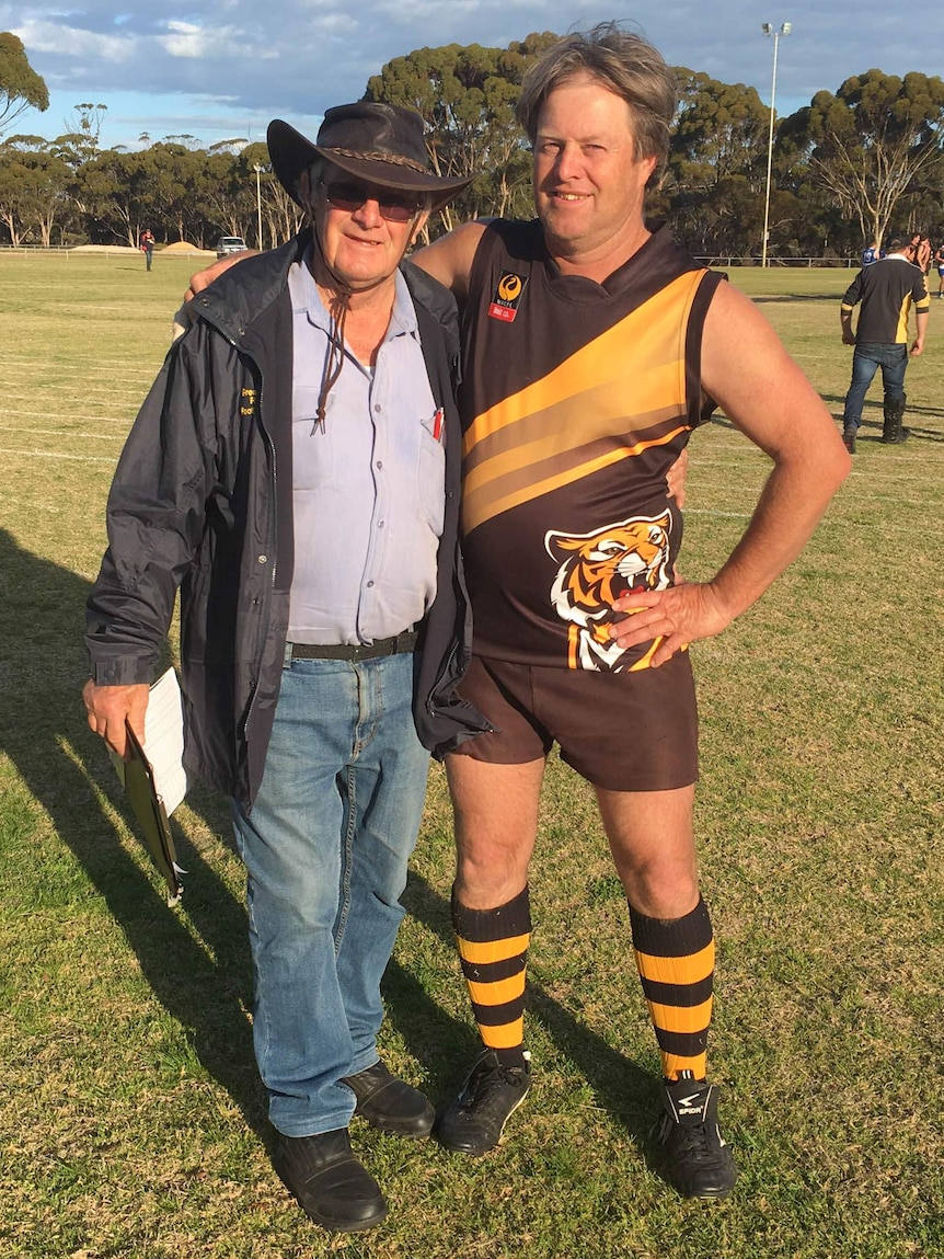 Two men stand on a football field, one of them wearing a black an gold playing strip.