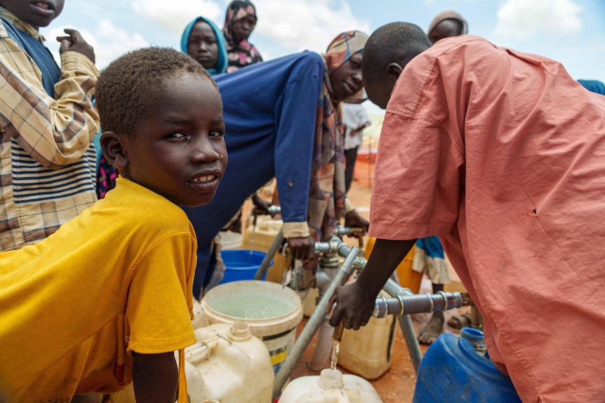A boy wearing a yellow shirt smiles as he leans of a bucket surrounded by people filling buckets with water