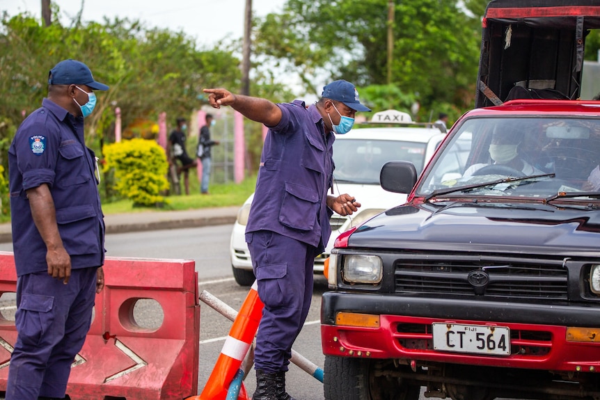 Two men in blue uniforms and surgical masks talk to the driver of a car.
