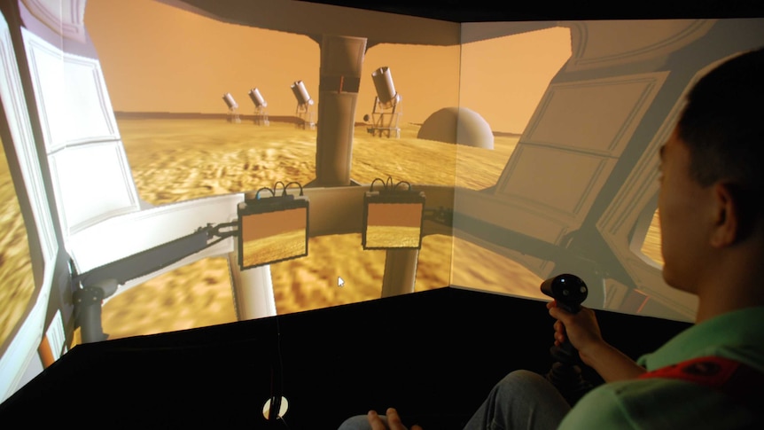 A man sits in a simulator, driving a vehicle on mars.