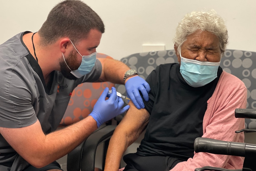 A male health worker vaccinates an Indigenous woman who is squeezing her eyes shut.