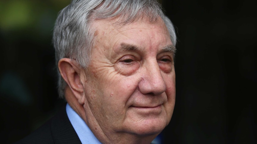 Profile of an older man with greying hair in a suit smiling and speaking to journalists