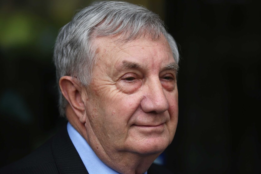 Profile of an older man with greying hair in a suit smiling and speaking to journalists