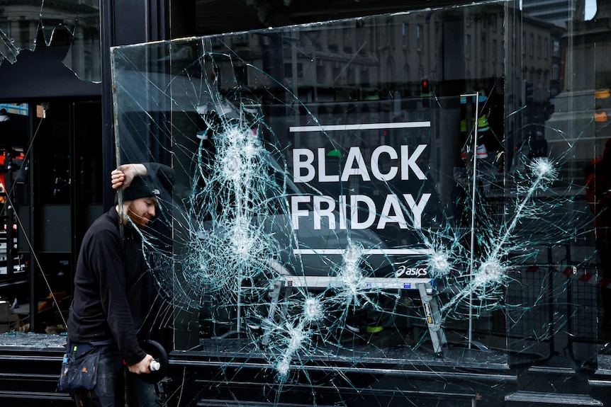 A man in a black beanie stands beside a cracked window saying "Black Friday"