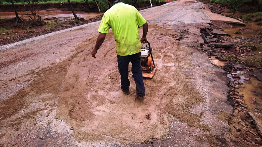 A worker with back to camera repairs a broken road with a compactor.