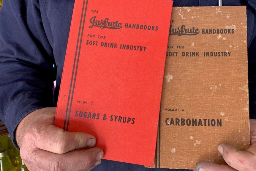 A man holds two 'handbooks', one red, one brown, full of recipes for soft drinks.