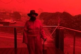 A woman in protective gear stands on a verandah against a bright red sky.