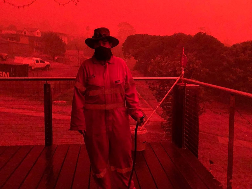 A woman in protective gear stands on a verandah against a bright red sky.
