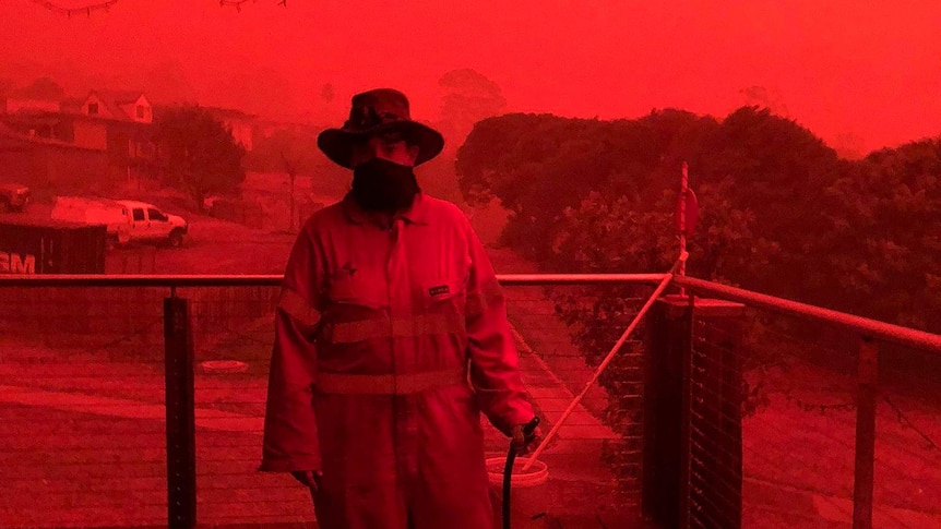 A woman in protective gear stands on a verandah against a bright red sky.