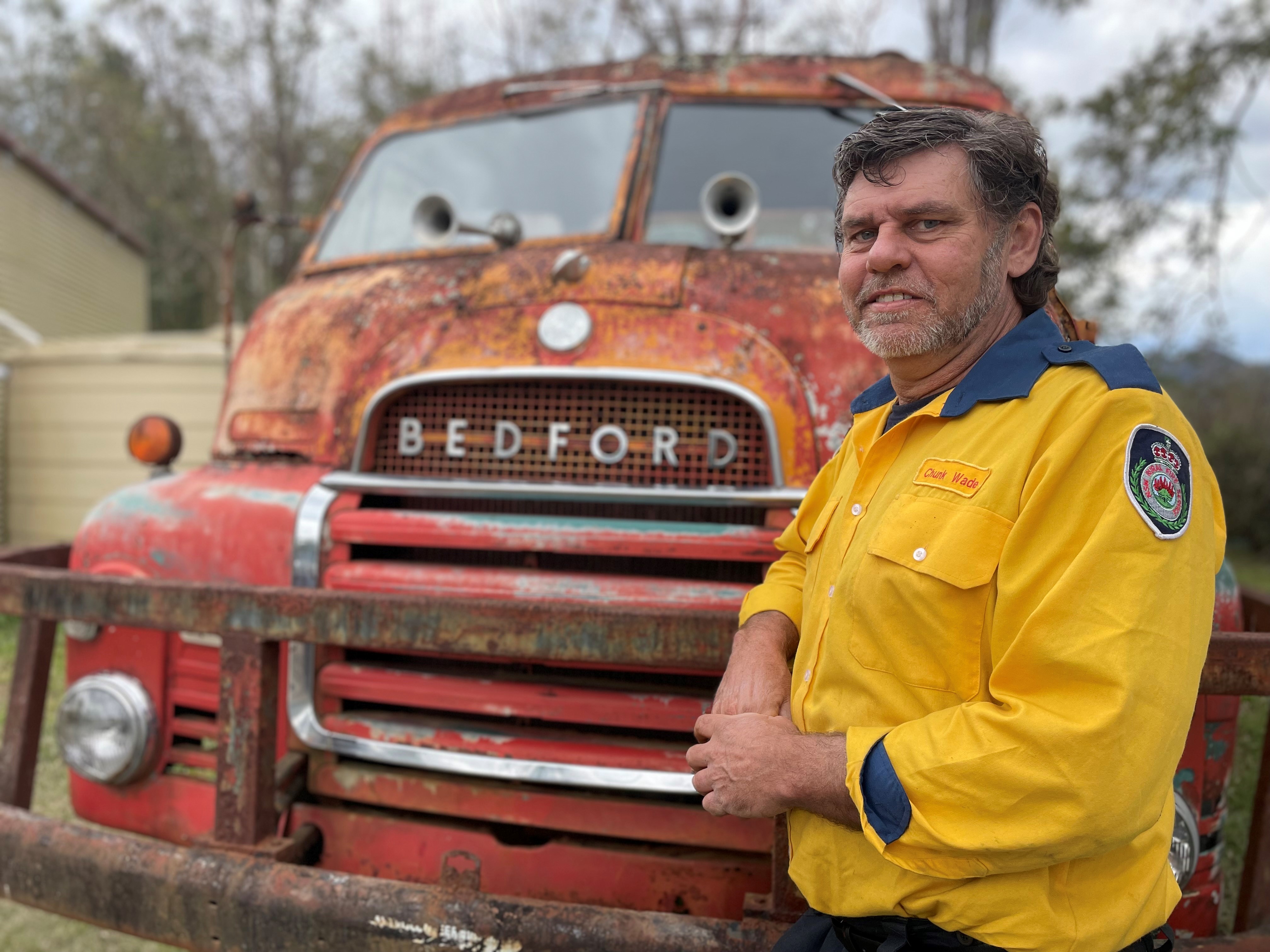 A firefighter leans on the front of an old rusty fire truck.