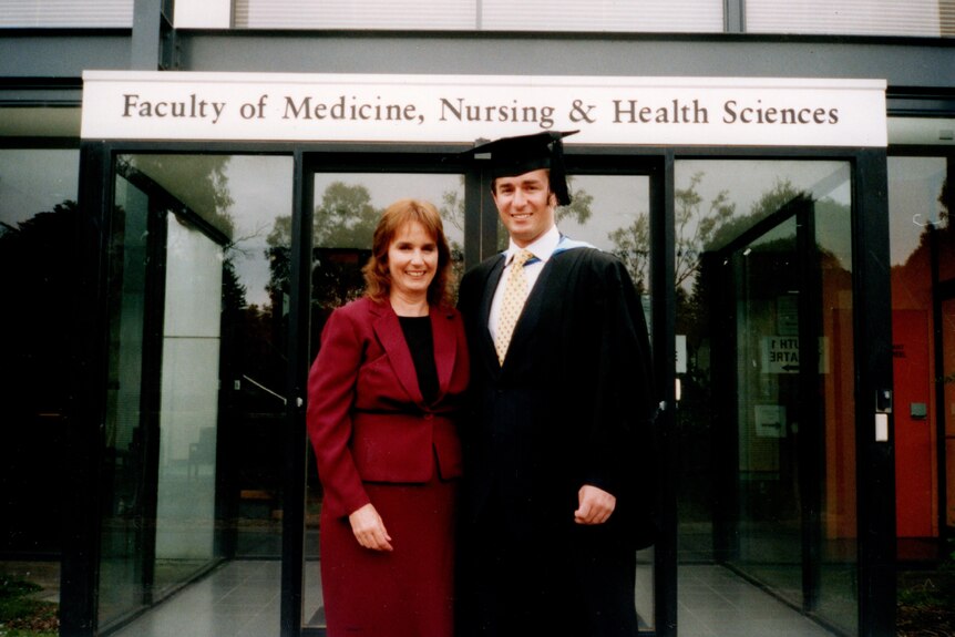 A smiling man in a graduation gown and hat standing with his mother