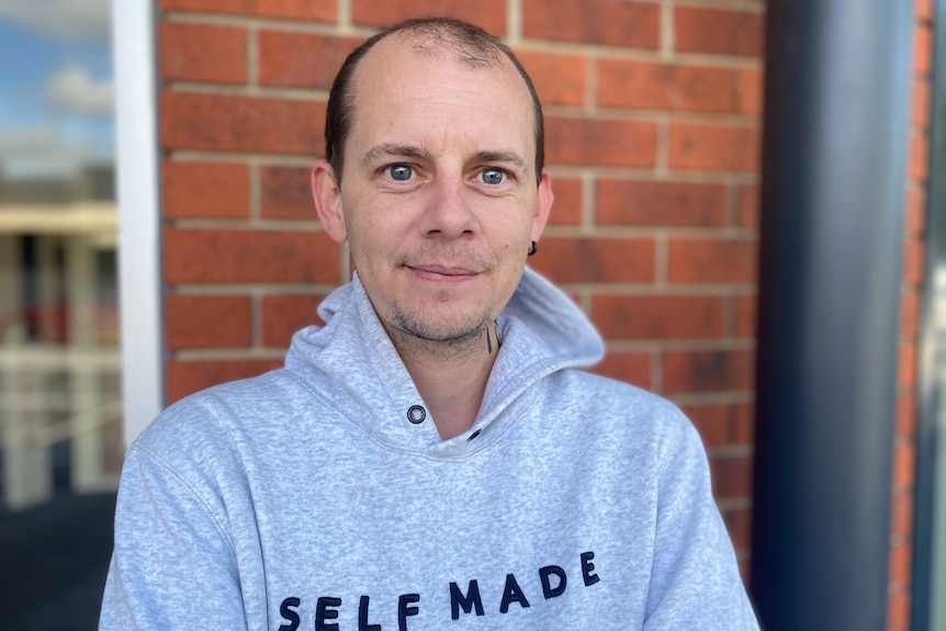 Richard Weeding stands in front of a red brick wall wearing a grey jumper