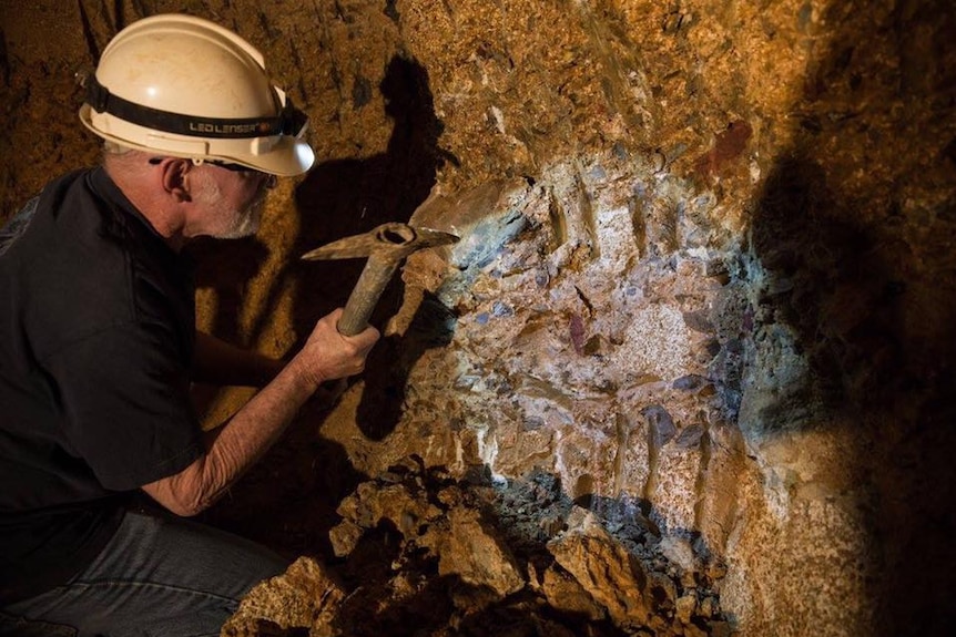 A man wearing a hard hat with a torch on it hammers at wall in an underground mine and shines a light on gem stones in the wall.