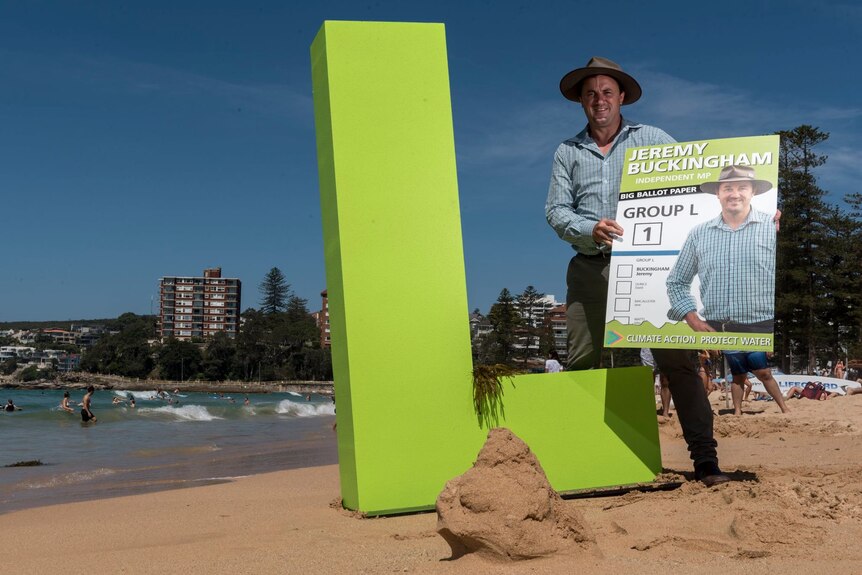 a man standing on the sand at a beach holding a large copy of a voting card 