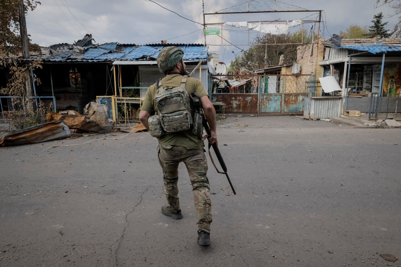 A soldier in fatigues, holding a rifle, runs towards a damaged building.