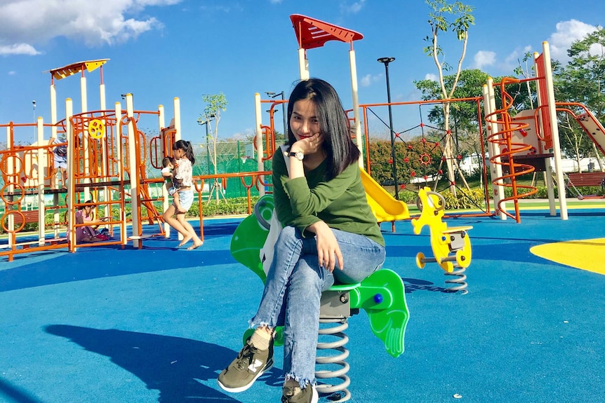 A woman sits on playground equipment, with a young child and baby in the background, a place to make new friends.