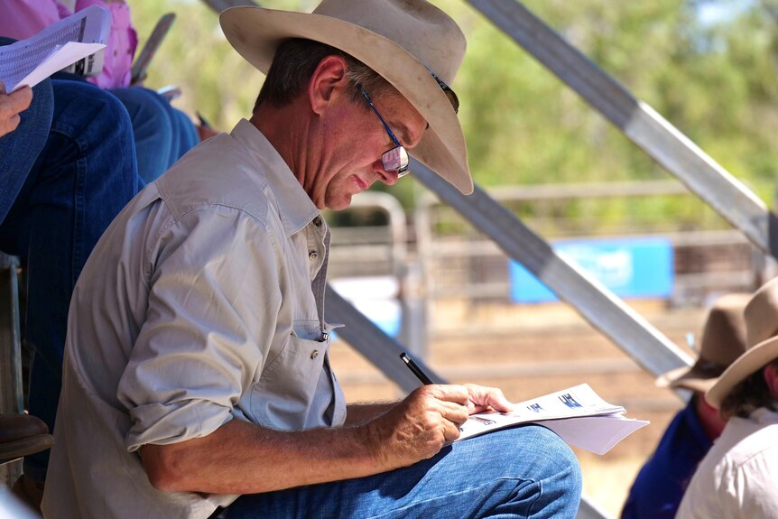 A man wearing reading glasses and a straw hat writing in a booklet on his lap