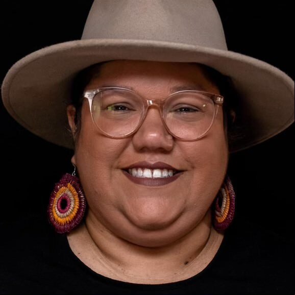 A smiling woman with glasses wears khaki Akubra style hat, background behind her is black.