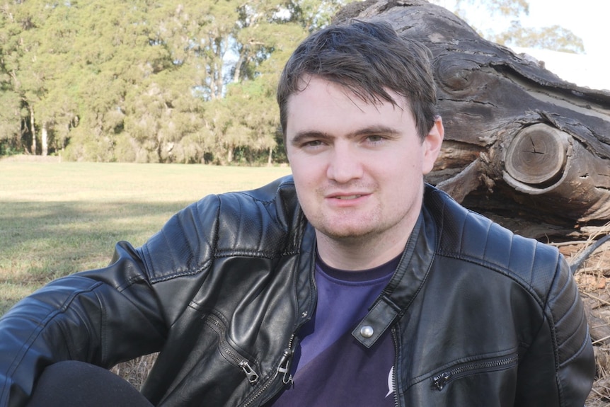 Young man wearing leather jacket sits under tree.