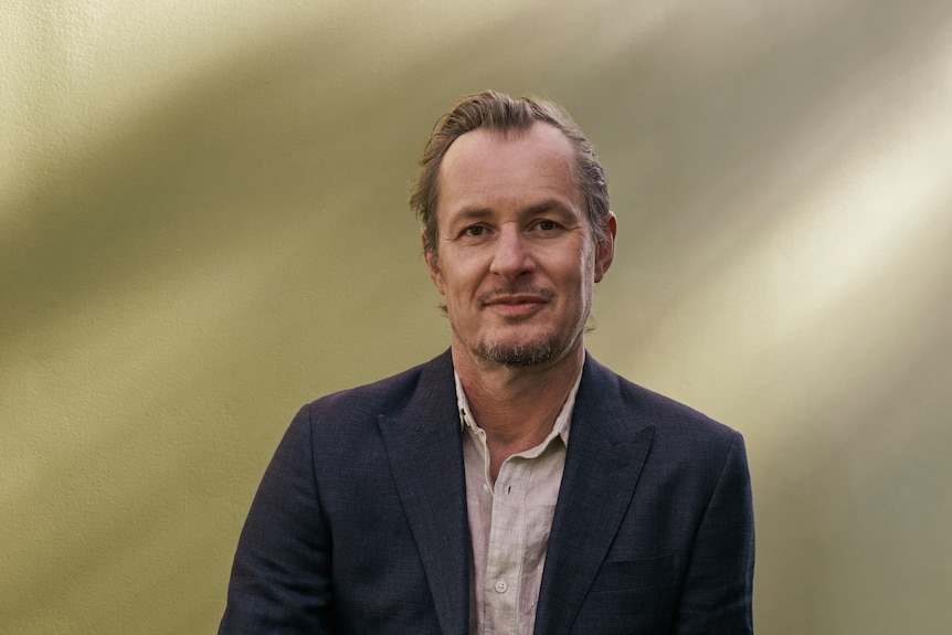 Richard Tognetti, a white man in his 60s, wears a black suit jacket over a cream linen shirt and leans against a taupe backdrop.