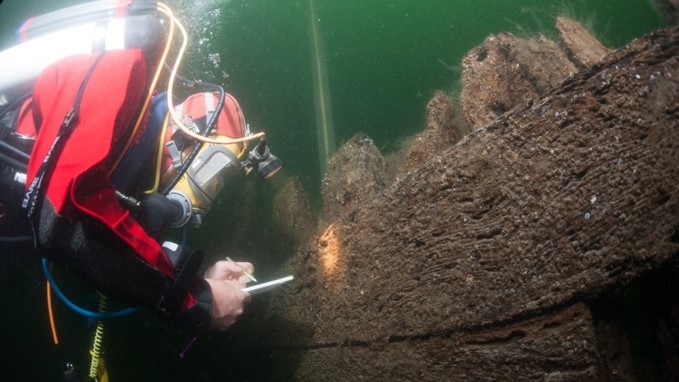 Underwater, you see a diver in a bright red watersuit and oxygen tank take notes next to brown timber.