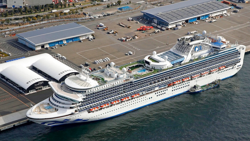 A large white cruise ship is pictured from above docked at a port with lots of empty car parking spaces.