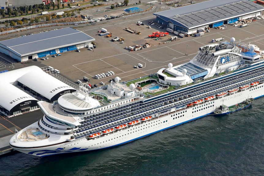 A large white cruise ship is pictured from above docked at a port with lots of empty car parking spaces.