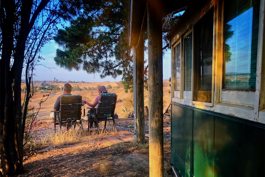 Two adults sit outside in garden chairs, their backs to the camera, looking over grassy paddocks bathed in golden light.