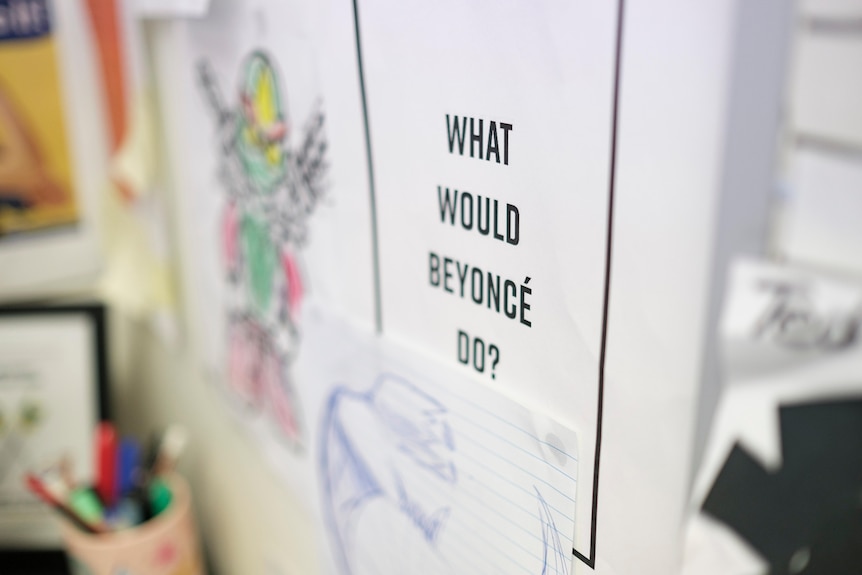 A white A4 piece of paper says What Would Beyonce Do? on it and is pinned to the wall of the office next to children's drawings.