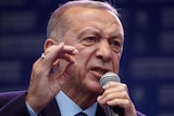 An older Turkish man in a suit yells into a microphone.